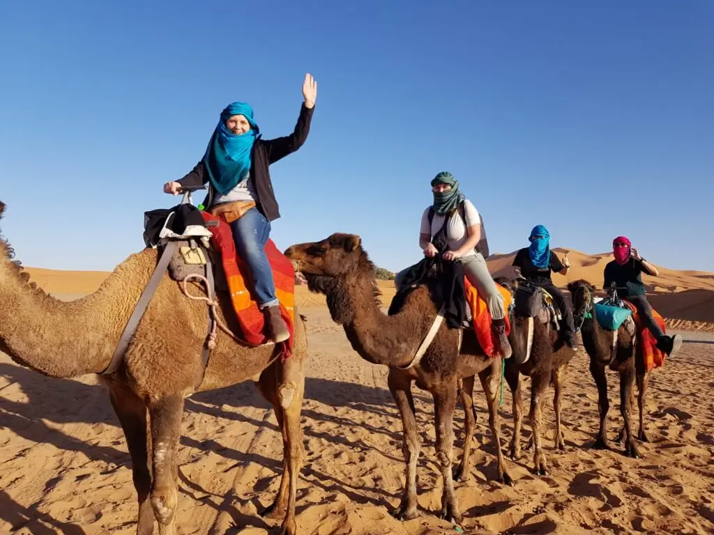 Tourists riding camels in the Moroccan desert, wearing colorful scarves and enjoying the desert scenery.